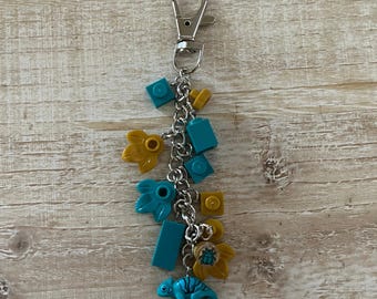 May include: A silver-tone keychain with a lobster clasp, adorned with a chain and various charms. The charms include teal and yellow building blocks, leaf shapes, and a small blue lizard figurine. The keychain is a decorative accessory.