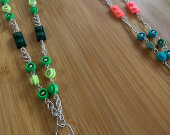 May include: Two lanyards constructed from silver-toned metal chains featuring integrated plastic building blocks and circular beads. One lanyard displays shades of green, while the other incorporates pink and blue elements, both ending in a metal clasp.