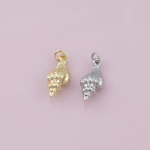 May include: Two seashell charms, one gold and one silver, sit against a soft pink backdrop. Each charm has a small loop at the top for attaching to a chain or bracelet. The charms are detailed with textured ridges, resembling miniature conch shells.