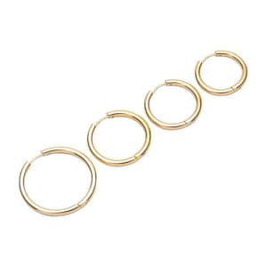Könnte beinhalten: Vier goldfarbene Creolen in verschiedenen Größen sind diagonal auf weißem Hintergrund angeordnet. Die Ohrringe haben ein schlichtes, poliertes Design mit glatter, abgerundeter Oberfläche. Die Creolen sind mit einem Klappverschluss verschlossen.