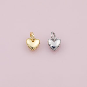 Peut inclure: Deux pendentifs en forme de cœur. L'un est doré, l'autre argenté. Chaque pendentif possède une petite boucle en haut pour l'attacher à une chaîne ou un bracelet. Les pendentifs sont présentés sur un fond rose clair.