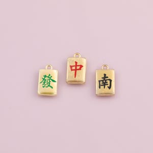 May include: Three gold-colored Mahjong tile charms with Chinese characters in red, green, and black. Each charm has a small loop at the top for attaching to jewelry or other crafts. The background is a soft pink.