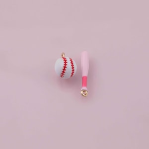 May include: A baseball and bat charm set. The baseball is white with red stitching. The bat is light pink with a darker pink bottom. Both charms have gold-coloured loops for attaching to jewellery or other crafts.