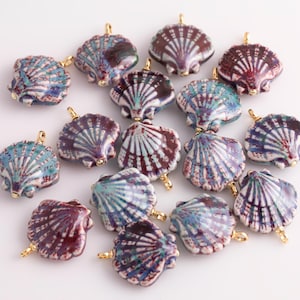 以下が含まれることがあります： 青、紫、白の色合いのセラミック製の貝殻ペンダントのコレクション。金色のループが付いています。白い表面に配置されています。ジュエリー作りやクラフトプロジェクトに最適です。