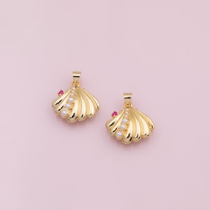 May include: Two gold-colored seashell pendants with a small red gem and a row of white pearls. The pendants have a loop at the top for a chain. The background is a solid light pink.