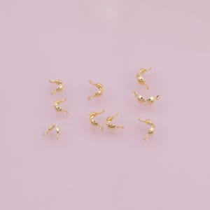 Può includere: Una collezione di piccoli elementi di bigiotteria dorati, probabilmente terminali a crimpare, sparsi su una superficie rosa chiaro. Ogni pezzo ha una forma di perla arrotondata con un'estremità curva e aperta, progettata per fissare i componenti dei gioielli.