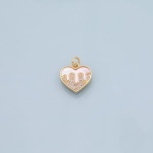 Puede incluir: Colgante dorado en forma de corazón con un diseño blanco ondulado y una sección de cristales transparentes. El colgante tiene un pequeño lazo en la parte superior para sujetarlo a una cadena o pulsera. El fondo es de color azul claro.