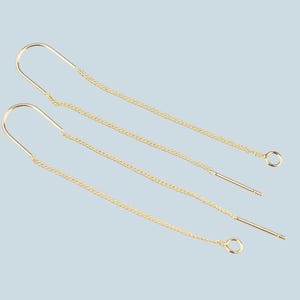 May include: A pair of delicate gold threader earrings. Each earring features a curved top, a chain, and a straight post. The earrings are displayed against a light blue background, showcasing their elegant design.