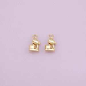 May include: Two identical gold-toned metal charms shaped like stand mixers. Each charm features a rounded top, a central arm, and a cylindrical base, with a small circular jump ring attached to the top for hanging or connecting to jewellery pieces.