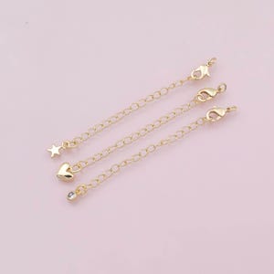 May include: Three gold-colored chain extenders with lobster claw clasps. One extender has a star charm, another a heart charm, and the third a small clear crystal charm. The extenders are on a light pink background.