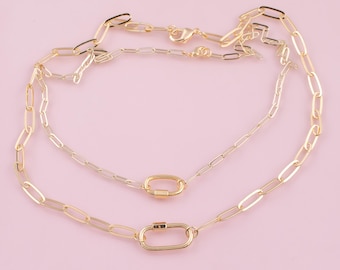 May include: Two gold-colored chain necklaces with oval links and a lobster clasp. The necklaces are layered, with one featuring a larger oval clasp. The background is a solid light pink.