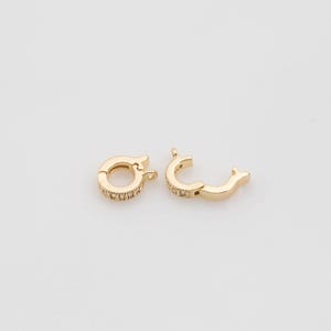 Könnte beinhalten: Zwei goldfarbene Schmuckverschlüsse, einer offen und einer geschlossen, werden vor einem weißen Hintergrund gezeigt. Die Verschlüsse haben eine runde Form mit kleinen, klaren Steinen an den Rändern. Der offene Verschluss ist gebogen, und beide haben kleine Schlaufen zum Befestigen.