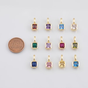 K&ouml;nnte beinhalten: Eine Sammlung goldfarbener quadratischer Anh&auml;nger mit verschiedenfarbigen Edelsteinen, darunter Rot, Lila, Blau und Gr&uuml;n. Eine M&uuml;nze liegt neben den Anh&auml;ngern zum Gr&ouml;&szlig;envergleich. Die Anh&auml;nger haben oben eine kleine Schlaufe zum Befestigen.