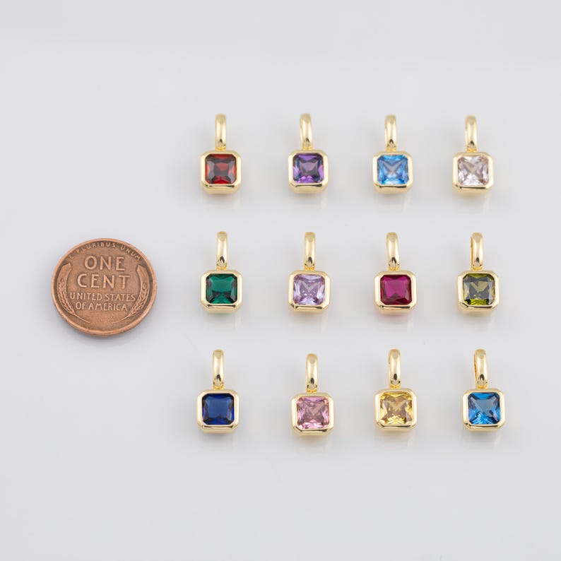 K&ouml;nnte beinhalten: Eine Sammlung goldfarbener quadratischer Anh&auml;nger mit verschiedenfarbigen Edelsteinen, darunter Rot, Lila, Blau und Gr&uuml;n. Eine M&uuml;nze liegt neben den Anh&auml;ngern zum Gr&ouml;&szlig;envergleich. Die Anh&auml;nger haben oben eine kleine Schlaufe zum Befestigen.