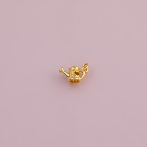 May include: A small, gold-colored watering can charm with a heart-shaped cutout and the words "planted in bloom." The charm has a loop for attaching to a necklace or bracelet. The watering can has a spout and a handle.