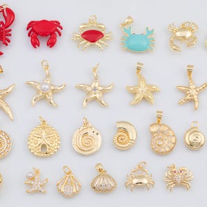 Op de afbeelding: Een verzameling goudkleurige en rode hangers met zeethema's, waaronder zeesterren, krabben en schelpen. Sommige hangers hebben parels of turkooizen accenten. De hangers zijn ontworpen voor het maken van sieraden.