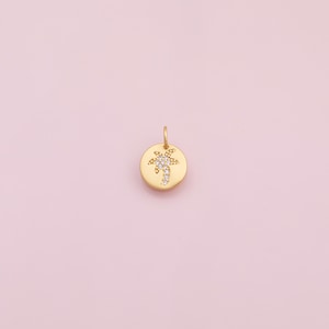 May include: A gold-colored pendant with a small loop at the top. The pendant is round with a palm tree design made of small, clear stones. The background is a soft pink.