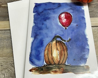 Peut inclure: Carte de vœux à l'aquarelle représentant une citrouille et un ballon rouge sur fond bleu. Une petite figurine de citrouille orange sur le thème d'Halloween avec des feuilles vertes et un visage effrayant est en arrière-plan. La carte est accompagnée d'une enveloppe blanche.