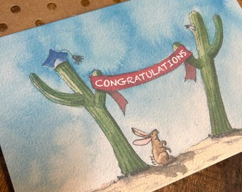 Peut inclure: Carte de félicitations de fin d'études à l'aquarelle avec deux cactus tenant une bannière rouge avec l'inscription "Félicitations". Un chapeau de remise de diplôme est sur un cactus et un lapin en dessous.