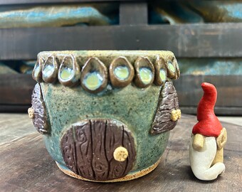 Peut inclure: Un petit pot en céramique vert en forme de maison avec une porte brune et des accents blancs. Une petite figurine de gnome avec un chapeau rouge se tient à côté du pot.