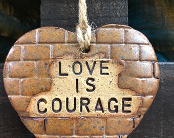 Peut inclure: Une décoration murale en céramique en forme de cœur avec un motif de briques texturé dans des tons marron et beige. Le centre présente le texte noir en relief LOVE IS COURAGE. Une ficelle de jute naturelle passe par un trou en haut.