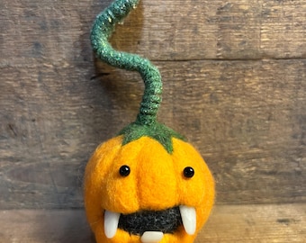 Peut inclure: Une figurine de citrouille orange en laine feutrée avec une tige torsadée vert foncé à paillettes. La citrouille présente deux petits yeux en perles noires, une zone buccale gris foncé et trois protubérances blanches pointues en forme de dents.