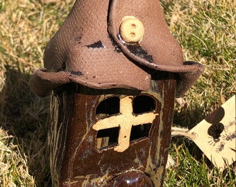 Peut inclure: Une maison de fée en céramique faite à la main, avec une finition marron texturée. La maison a un toit pointu fixé avec un bouton, une fenêtre en forme de croix et une porte en forme de cœur. La maison est posée sur de l'herbe verte.