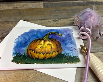 Peut inclure: Peinture à l'aquarelle d'une citrouille sculptée avec une tige verte, sur fond bleu et violet. La citrouille est orange avec un visage sculpté. La peinture est sur une carte blanche, avec une baguette décorative en arrière-plan.