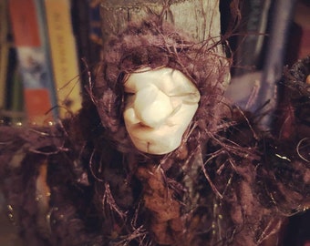 Peut inclure: Une poupée artisanale au visage sculpté pâle, dotée d'un nez proéminent et d'yeux fermés, portant une capuche et un vêtement en fil texturé brun foncé et duveteux qui recouvre la tête et les épaules, placée contre une structure en bois.
