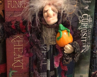 Peut inclure: Une figurine artisanale avec un visage sculpté pâle au nez proéminent et des cheveux blancs ébouriffés, fixée sur un torse en bois texturé. Le personnage porte un tissu sombre effiloché et tient une petite citrouille orange à tige verte.