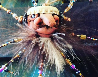 Peut inclure: Un ornement artisanal composé d'un visage en argile sculpté au nez proéminent, coiffé d'une capsule de bouteille beige. La figurine possède une barbe en plumes brunes et des membres rayonnants faits de perles en verre colorées.