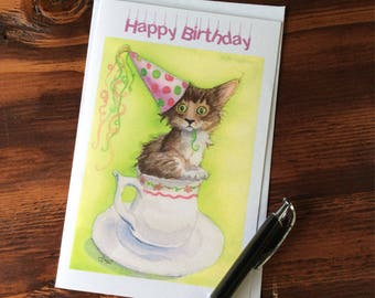 Peut inclure: Une carte d'anniversaire illustrant un chat en aquarelle portant un chapeau de fête et assis dans une tasse à thé. La carte porte le texte "Happy Birthday" imprimé dessus.