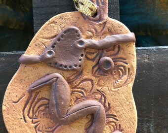 Peut inclure: Un ornement en céramique beige à la finition mouchetée, présentant une figurine en argile brun foncé en relief en position agenouillée et un accent en forme de cœur. La surface est ornée de motifs d'araignées gravés et suspendue par un ruban doré.