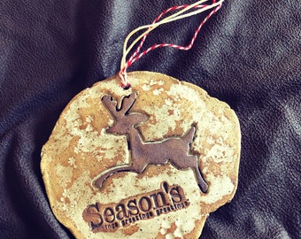 Peut inclure: Ornement de Noël en céramique avec une silhouette de renne et l'inscription "Season's greetings". L'ornement est beige avec des détails marron et une ficelle rouge et blanche. Le renne saute.
