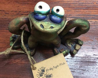 Peut inclure: Une figurine de grenouille sculptée verte avec de grands yeux blancs, des paupières bleu foncé et une finition métallique. Une étiquette en papier brun est attachée avec de la ficelle, portant le texte Grubby Angels Studio et trois empreintes.