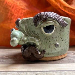 May include: A handmade ceramic mug with a whimsical face design. The mug features a green speckled glaze, a large eye, and a sculpted mustache. The details are in brown, and the mug has a natural clay base.