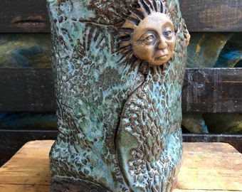 Peut inclure: Un vase en céramique fait à la main avec une surface texturée et mouchetée dans des tons de vert d'eau et de marron. Le vase présente un visage de soleil sculpté avec des rayons. Le design a une esthétique rustique et terreuse.