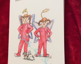 Peut inclure: Une carte de vœux blanche avec une illustration de deux enfants en pyjama rouge, portant des ailes bleues et des auréoles, l'air grincheux. Le texte indique : Humbug ~ Grumblebug. There's no Humbug in Christmas! Un petit chien porte des oreilles de lapin.