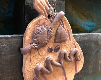 Peut inclure: Une décoration en argile beige tachetée en forme de citrouille avec une tige et des traits de visage marron foncé. La citrouille présente un œil triangulaire, un petit œil circulaire et une bouche ondulée en relief marron foncé.