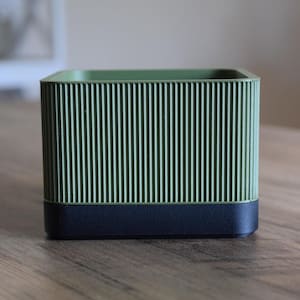 Könnte beinhalten: Ein grüner und schwarzer rechteckiger Behälter mit vertikalen Rippendetails. Der obere Teil ist hellgrün, während die Basis schwarz ist. Der Behälter steht auf einer Holzoberfläche.