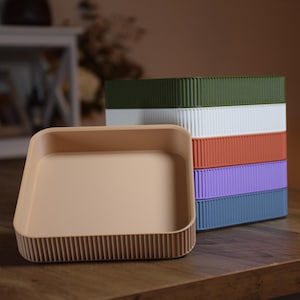May include: A selection of colourful, ribbed storage containers in green, white, orange, purple, and blue. A beige tray with a ribbed base is in front of the stack, on a wooden surface.