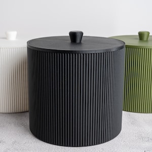 Puede incluir: Tres recipientes cilíndricos con tapa, cada uno con una textura acanalada vertical. Los recipientes son de color blanco, negro y verde oliva, y cada tapa está rematada con un pequeño pomo a juego para facilitar su apertura y acceso.