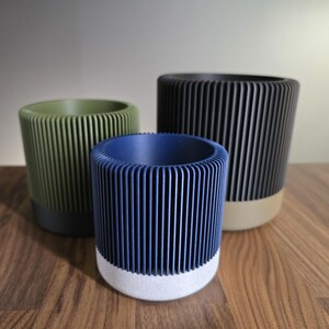 May include: Three cylindrical planters in olive green, navy blue, and black, each with vertical ribbed detailing. The planters have contrasting base colours: dark grey, white, and beige. They are arranged on a wooden surface.
