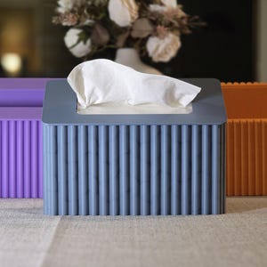 Könnte beinhalten: Drei rechteckige Taschentuchboxen in Lila, Blau und Orange. Die Boxen haben ein geripptes Design und sind auf einem Tisch mit einem neutralfarbenen Tischläufer angeordnet. Im Hintergrund steht eine Vase mit weißen Blumen.