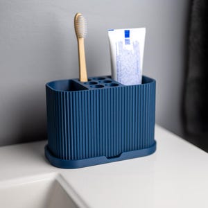 May include: A dark blue, oval-shaped bathroom organizer with a vertical ribbed texture. The container features multiple compartments of varying sizes, currently holding a wooden toothbrush and a tube of toothpaste in an upright position.