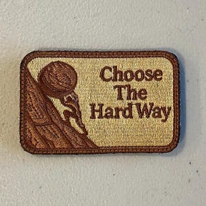 May include: A rectangular patch with a tan background and brown border. The patch features a brown illustration of a person climbing a mountain and the text "Choose The Hard Way" in brown.