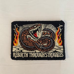 May include: Black embroidered patch with a snake wrapped around itself with flames on either side. The text "Rebirth Through Struggles" is below the snake.