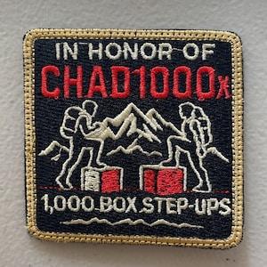 May include: Embroidered patch with a black background and gold trim. The text "IN HONOR OF CHAD1000x" is in red and white. Two figures climb mountains and boxes. The text "1,000 BOX STEP-UPS" is in white.