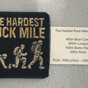 May include: Black square patch with gold embroidered text "THE HARDEST RUCK MILE" and illustrations of a person in a military backpack in different stages of movement. A white card lists the workout details.