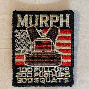 May include: A black and white embroidered patch with the word "MURPH" in white lettering. The patch features a stylized American flag with a brown and white vest over it. The text "100 PULL-UPS 200 PUSH-UPS 300 SQUATS" is embroidered in white lettering below the flag.